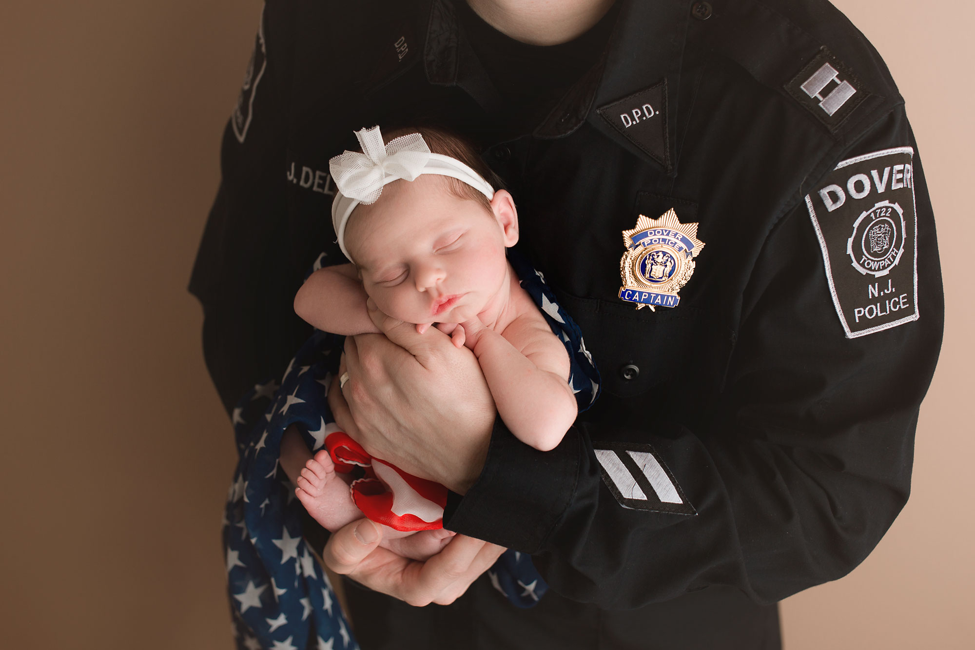 dad wearing a police officer uniform holding a newborn baby girl swaddled in red, white and blue scarf resembling an American flag. Morris county NJ newborn photographer , Yellow Lollipop Photography