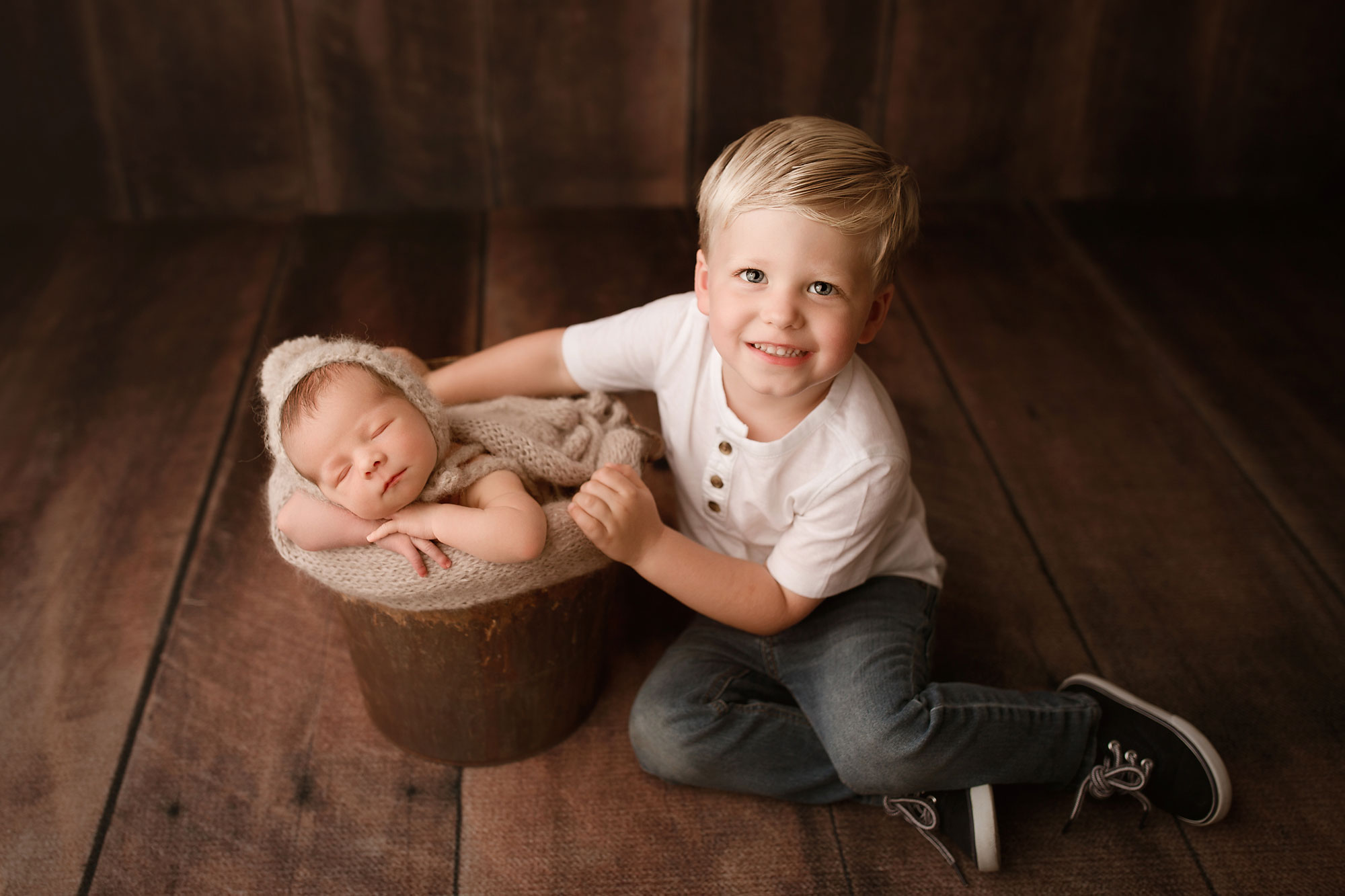 newborn and sibling photographer new jersey, little boy seated next to baby sleeping in rustic bucket on wood backdrop