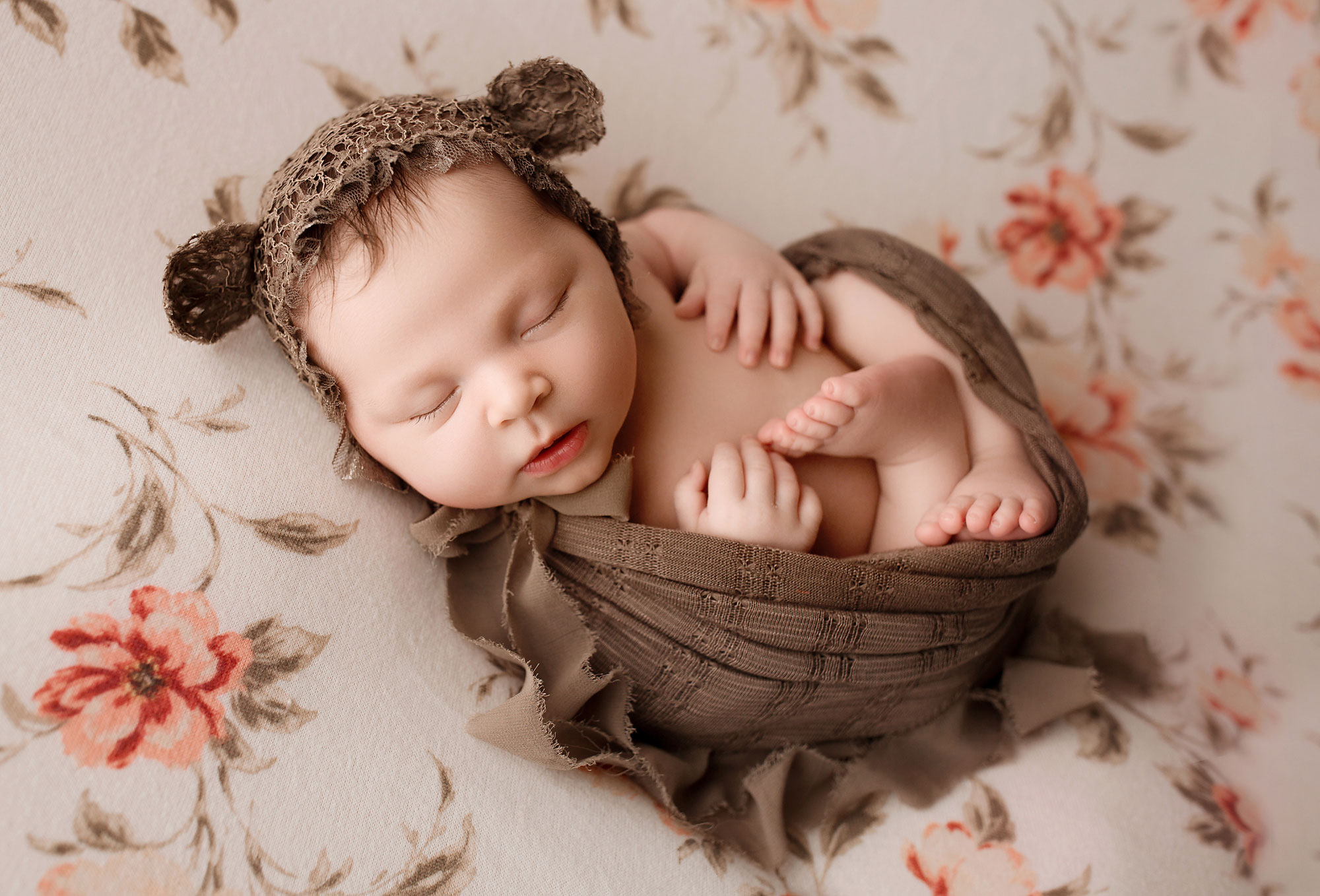professional newborn photography near me, baby girl in brown wrap and bear cap on floral backdrop