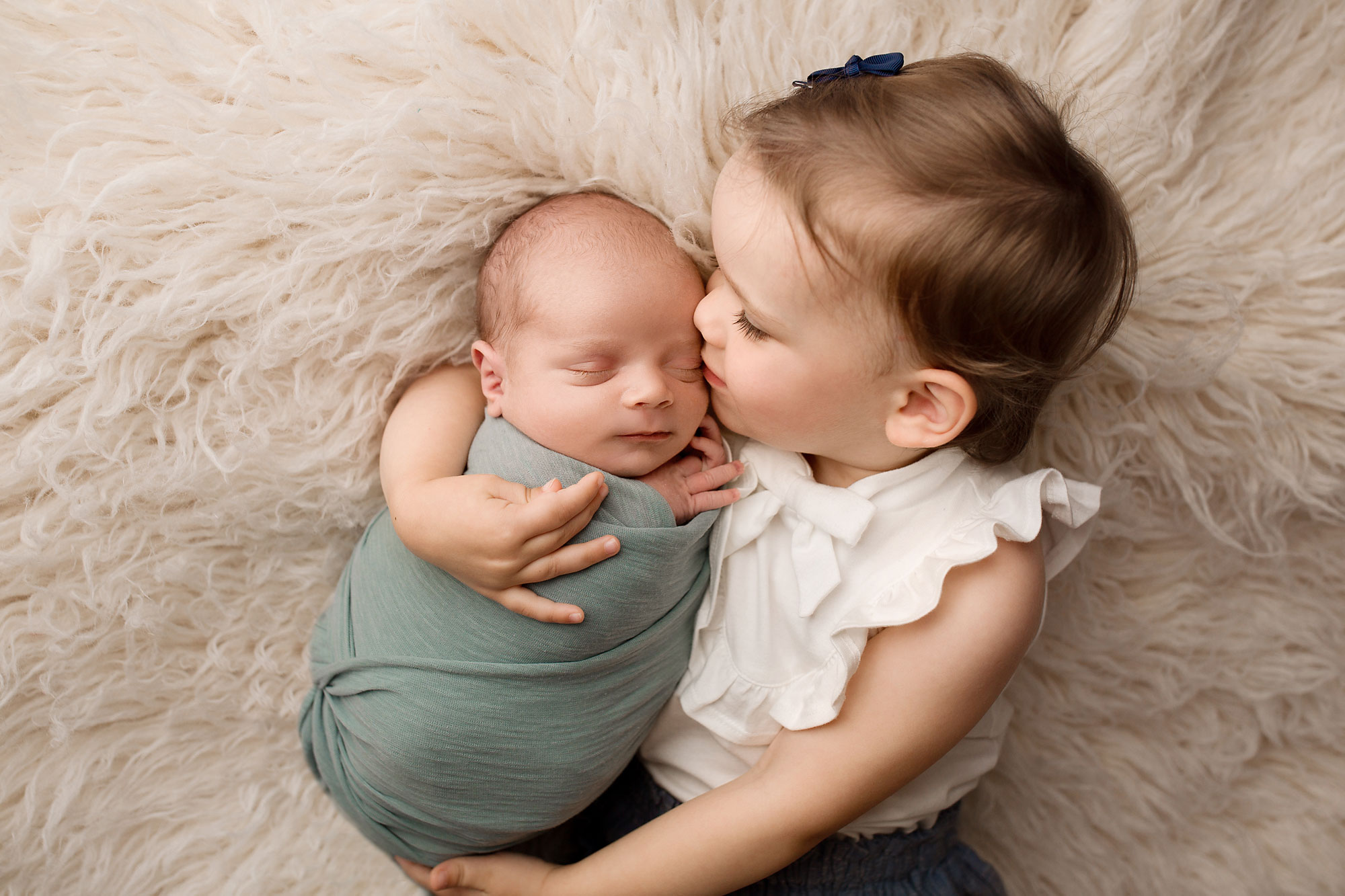 professional newborn and sibling photography, little girl snuggling swaddled baby against a soft fur background