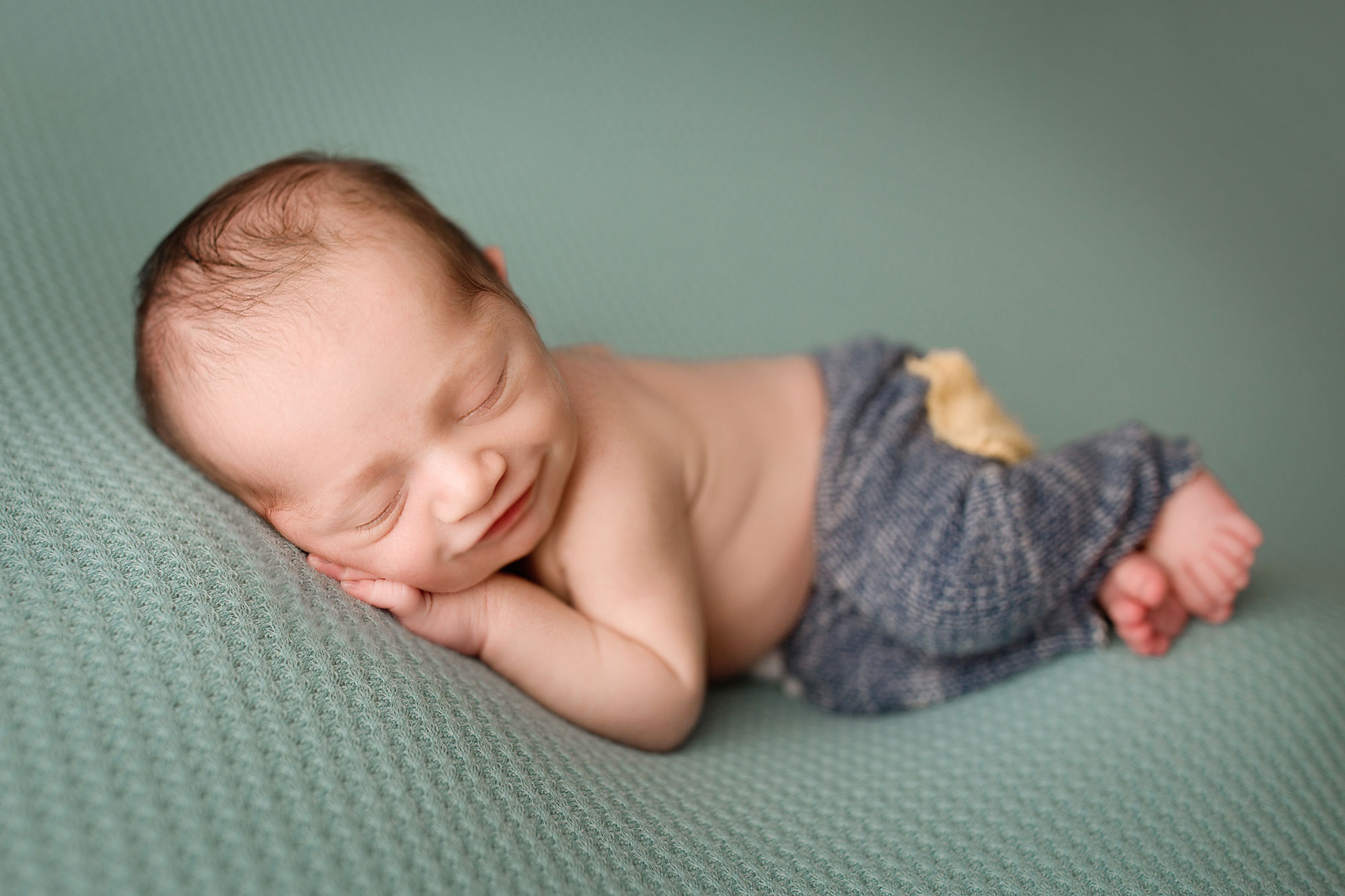 Branchburg NJ newborn pictures, smiling baby boy dressed in patched pants and asleep on teal background