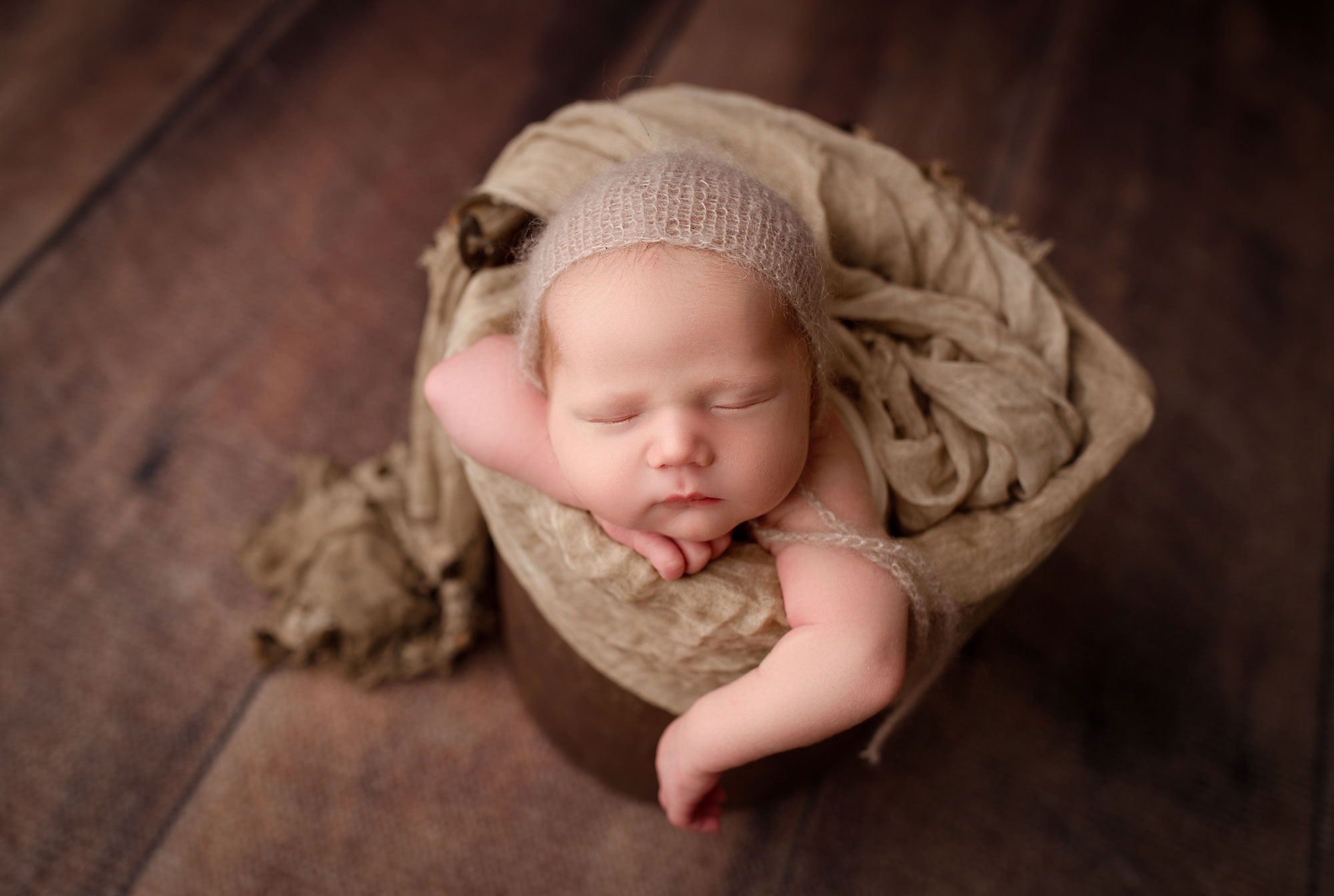 newborn pictures, baby boy in light brown wrap and cap sleeping in bucket against dark wood backdrop