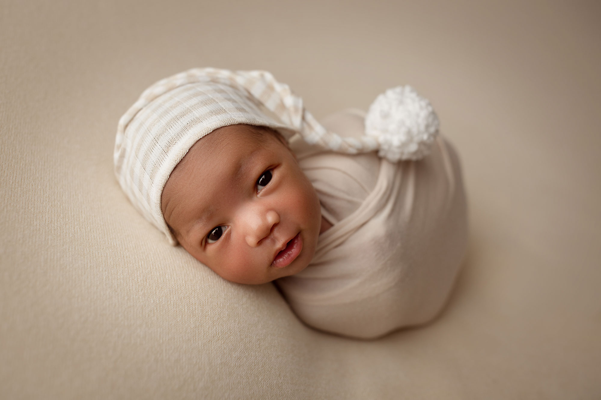 newborn pictures near me, swaddled baby boy in white hat looking at camera