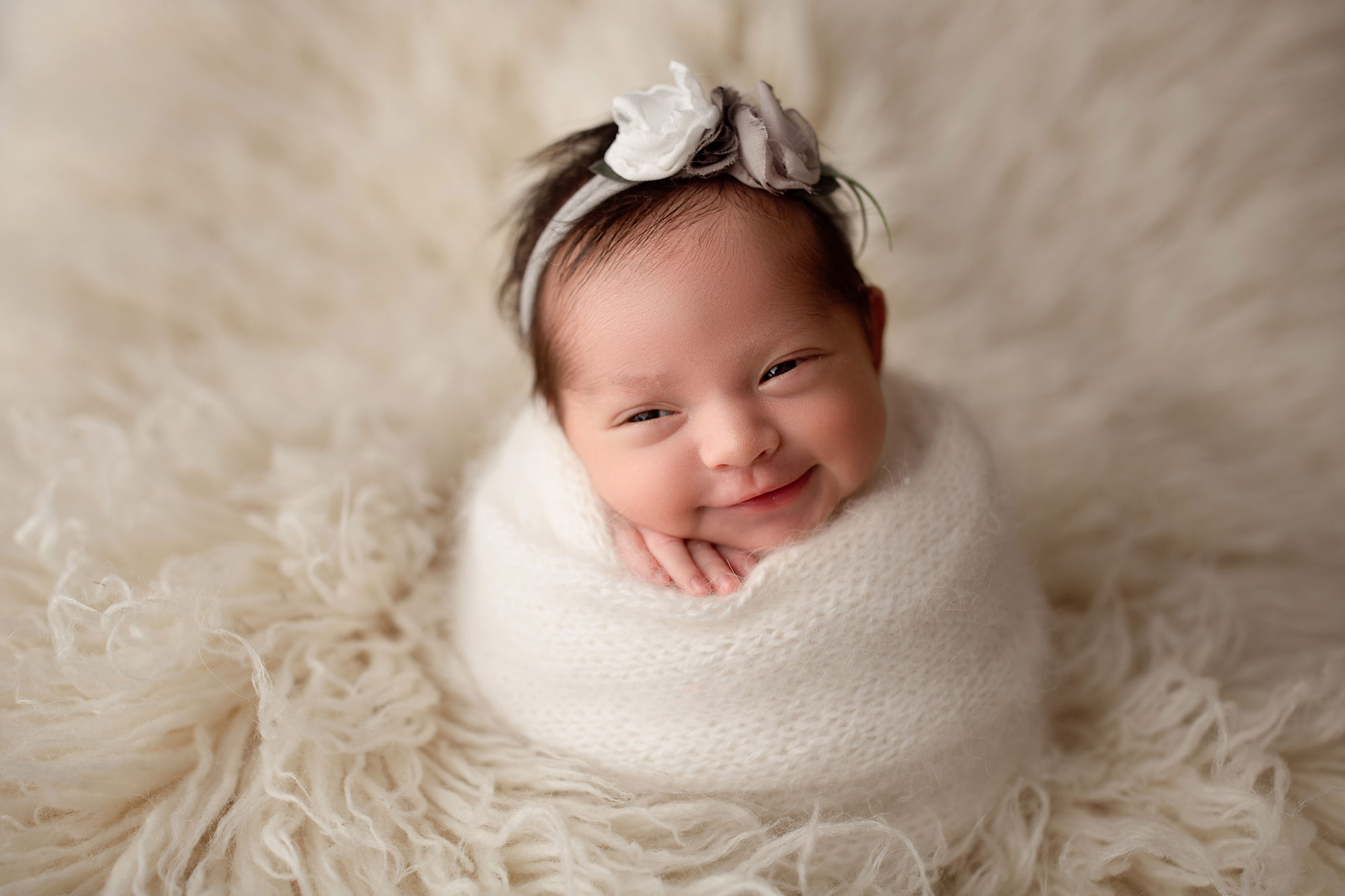 smiling baby girl swaddled in a white wrap on a cream flokati 